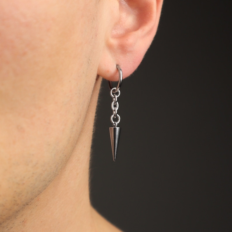 May include: A silver-colored earring with a chain and a cone-shaped pendant. The earring is attached to a hoop that is fastened to the ear. The earring is shown against a dark background.