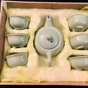 Pode incluir: Um conjunto de chá de cerâmica verde-pálido composto por um bule com tampa e seis pequenas chávenas a condizer. O conjunto está disposto dentro de uma caixa retangular forrada com tecido amarelo claro, com compartimentos moldados para cada peça.