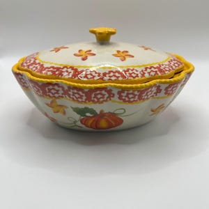 May include: A ceramic serving bowl with a matching lid, featuring a scalloped yellow rim and a decorative orange floral pattern. The exterior is adorned with a painted orange pumpkin and green leaves, topped with a yellow flower-shaped handle.