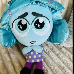 May include: A plush doll featuring a light blue head with large embroidered eyes, dark eyebrows, and teal hair with pink accents. The doll wears a purple shirt with yellow polka dots, purple pants, and black shoes, with arms crossed in front.