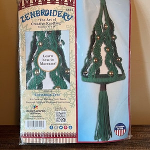 Könnte beinhalten: Ein Zenbroidery Makramee-Set für einen Weihnachtsbaum mit grüner Kordel, goldenen Perlen und einer Quaste. Die Verpackung zeigt den Text "The Art of Creative Knotting", "Finishes 20.32 cm x 60.96 cm" und "Learn how-to Macrame!" sowie einen QR-Code.