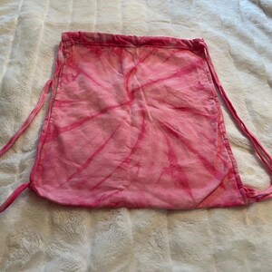 May include: A pink and white tie-dye drawstring backpack featuring a spiral pattern. The bag is constructed from fabric with matching pink cord straps that cinch the top opening closed, creating a gathered effect at the upper edge of the bag.
