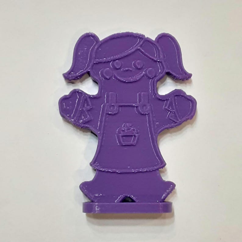 Pu&ograve; includere: Figurina di plastica viola di una ragazza con le trecce che indossa un vestito e una salopette. La ragazza tiene le braccia distese ai lati.