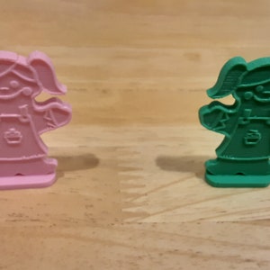 Pu&ograve; includere: Due figurine di plastica di ragazze con vestiti e codini. Una figurina &egrave; rosa e l'altra &egrave; verde.