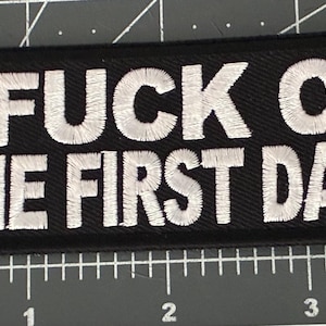 May include: Black rectangular patch with white embroidered text that reads "I FUCK ON THE FIRST DATE". The patch is on a black surface with a ruler in the background.