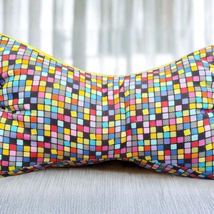 May include: A colorful, butterfly-shaped pillow with a vibrant patchwork design. The pillow features a repeating pattern of small squares in various colors, including blue, yellow, pink, and green, set against a black background. The edges are trimmed with yellow.