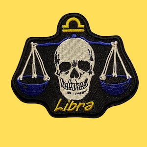 May include: Black and white embroidered patch of a skull with a scale balance and the word "Libra" in yellow. The scales are blue and white.