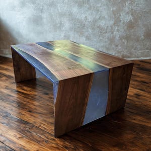 May include: A handcrafted wooden coffee table with a unique design. The table features a dark wood finish with a clear, resin-filled channel running through the center. The table is approximately 18 inches tall, 48 inches long, and 24 inches wide.