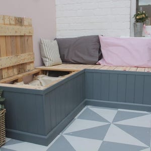 Peut inclure: Banc extérieur en forme de L avec un compartiment de rangement, peint en gris. Le banc est surmonté d'un couvercle et d'une assise en bois, avec des coussins gris et roses. Un panier en osier et une plante en pot sont au premier plan.