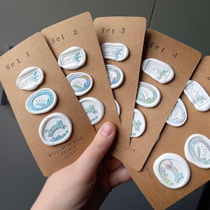 Op de afbeelding: Een hand houdt verschillende sets witte waszegels op bruin karton. Elke zegel heeft een uniek ontwerp met blauwe en groene accenten. De sets zijn gelabeld met "Set 1" tot "Set 4". De tekst "FULLMETALMILK PRINTS -ETSY-" is zichtbaar.