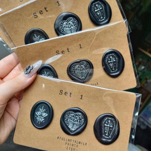 May include: Black wax seals with white designs, including a coffin, a heart with skeletons, and a moth. Each set is labelled "Set 1" on a brown card. The Etsy shop name "FULLMETALMILK PRINTS - ETSY -" is printed at the bottom.
