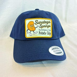 May include: Navy blue baseball cap with a patch featuring a smiling potato chip character and the text "Saratoga Springs The Birthplace of the Potato Chip." The cap has an adjustable strap and a small white tag.