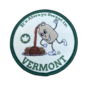 May include: A circular embroidered patch with a dark green border and white background. It features a cartoon jug pouring brown syrup onto a stack of pancakes. The text reads "It's Always Sweet in VERMONT" and includes a green maple leaf icon.