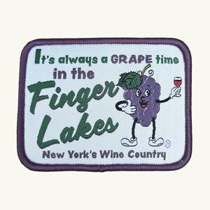 May include: A rectangular embroidered patch with a light blue background and a dark purple border. It features a cartoon purple grape character holding a wine glass, with the text: It's always a GRAPE time in the Finger Lakes New York's Wine Country.