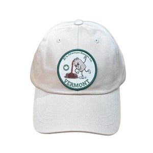 May include: A light beige baseball cap featuring a circular patch on the front. The patch has a dark green border and displays a cartoon maple syrup jug pouring syrup onto a stack of pancakes with the text It's Always Sweet in VERMONT.