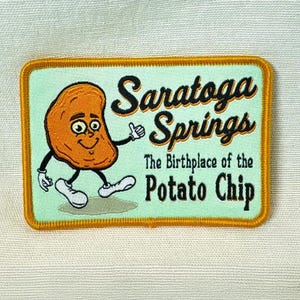 May include: Embroidered patch featuring a smiling potato chip character with arms and legs. The patch reads "Saratoga Springs" and "The Birthplace of the Potato Chip" on a light green background with a gold border.
