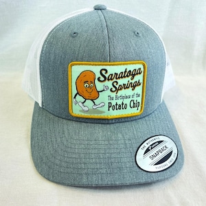 May include: A gray and white trucker hat with a patch that reads "Saratoga Springs The Birthplace of the Potato Chip." The patch features a cartoon potato character. The hat has a snapback closure.