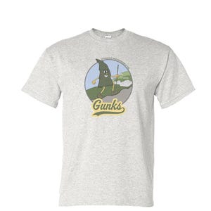 May include: Light gray t-shirt featuring a cartoon graphic of a tree-like character holding a stick, with the text "Gunks" in a vintage font. The graphic is set within a circular design with the phrase "It's always tree-mendous in the Gunks."
