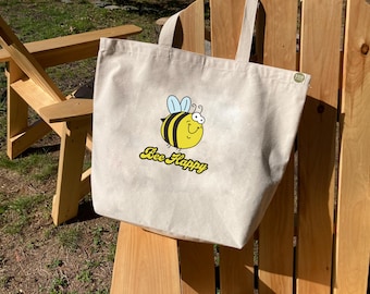 May include: A beige tote bag with a cartoon bee and the text "Bee Happy". The bag has a brown leather handle and a small brown tag that says "Eco Friendly".