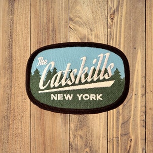 May include: An embroidered patch with the text "The Catskills NEW YORK" in white script. The patch features a light blue sky, green trees, and a dark brown border, set against a wooden background.