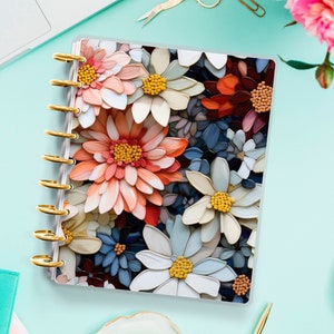 May include: A planner with a 3D floral design cover in peach, white, and blue. The planner has gold rings and is open. The background includes a laptop, stationery, and flowers. The planner's cover features a variety of flowers.