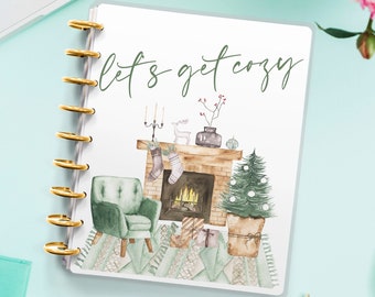 Può includere: Un'agenda bianca con una rilegatura a spirale dorata. La copertina presenta un'illustrazione ad acquerello di un camino con una poltrona verde, un albero di Natale e il testo "Let's get cozy".