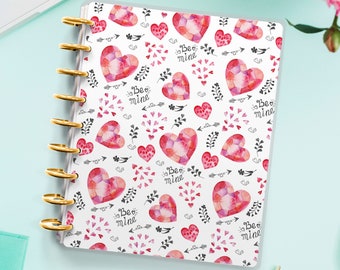 Op de afbeelding: Een witte planner met een transparante hoes met een herhalend patroon van roze aquarelhartjes met de tekst "Be mine" in zwart. De planner heeft gouden ringen en ligt op een lichtblauw oppervlak.