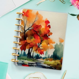 Puede incluir: Una agenda de plástico transparente con un diseño de acuarela de una escena de otoño con hojas naranjas, rojas y amarillas en un árbol. La agenda tiene anillos dorados y un fondo blanco.