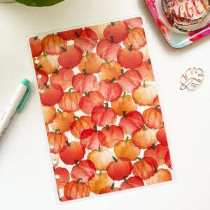 May include: A clear plastic sheet with a repeating pattern of watercolor pumpkins in shades of orange and red. The sheet is designed for use in a planner or journal.