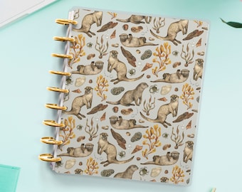 May include: A gray and white planner cover with a repeating pattern of otters, seaweed, and seashells. The otters are brown and white and are swimming in the water. The seaweed is green and brown and is swaying in the water. The seashells are white and are scattered on the ocean floor.