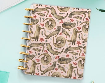 May include: A pink and white planner with a repeating pattern of brown otters in various poses. The otters are surrounded by small, pink and white flowers and leaves.