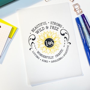 May include: White notebook with a black sunflower design and the text "I am beautiful, strong, wild & free, wonderfully chaotic, loving, kind, amazing, positive".