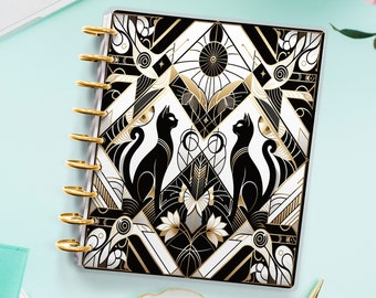 Könnte beinhalten: Ein schwarzer und goldener geometrischer Design-Planer mit zwei schwarzen Katzen, die sich gegenüberstehen. Der Planer hat eine transparente Abdeckung und eine goldene Spiralbindung.