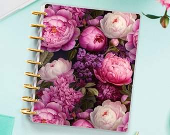 Op de afbeelding: Een notitieboek met een gouden spiraalbinding en een bloemenpatroon. De omslag heeft roze, witte en paarse pioenen en andere bloemen op een donkere achtergrond.