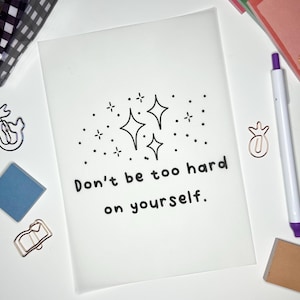 May include: A white card with black text that reads "Don't be too hard on yourself."  The text is surrounded by a black and white illustration of stars.