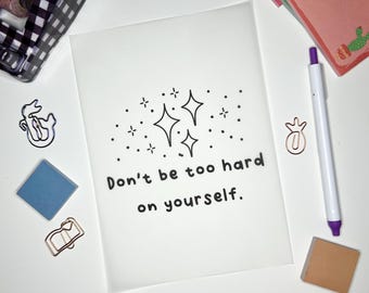 Può includere: Un biglietto bianco con testo nero che dice "Non essere troppo duro con te stesso." Il testo è circondato da un'illustrazione in bianco e nero di stelle.