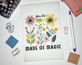Può includere: Un biglietto bianco con un disegno floreale e il testo "you are made of magic". I fiori sono rosa, gialli e verdi.