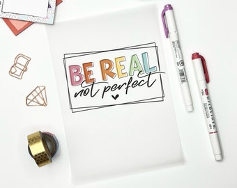 Può includere: Un foglio di carta bianco con la frase "BE REAL not perfect" scritta in colori arcobaleno. La frase è circondata da un contorno nero. Ci sono due pennarelli bianchi con tappi rosso e viola sul lato destro dell'immagine.