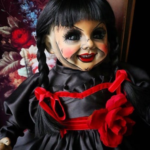 May include: A vintage doll with a porcelain face, black hair in braids, and a black dress with a red ribbon and rose accent. The doll has painted facial features, including red lips and rosy cheeks, and is posed against a floral background.