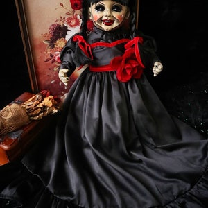 May include: A vintage-style doll with a porcelain face, black hair in pigtails, and a black dress with red accents. The doll has a painted smile and is posed in front of a floral print.