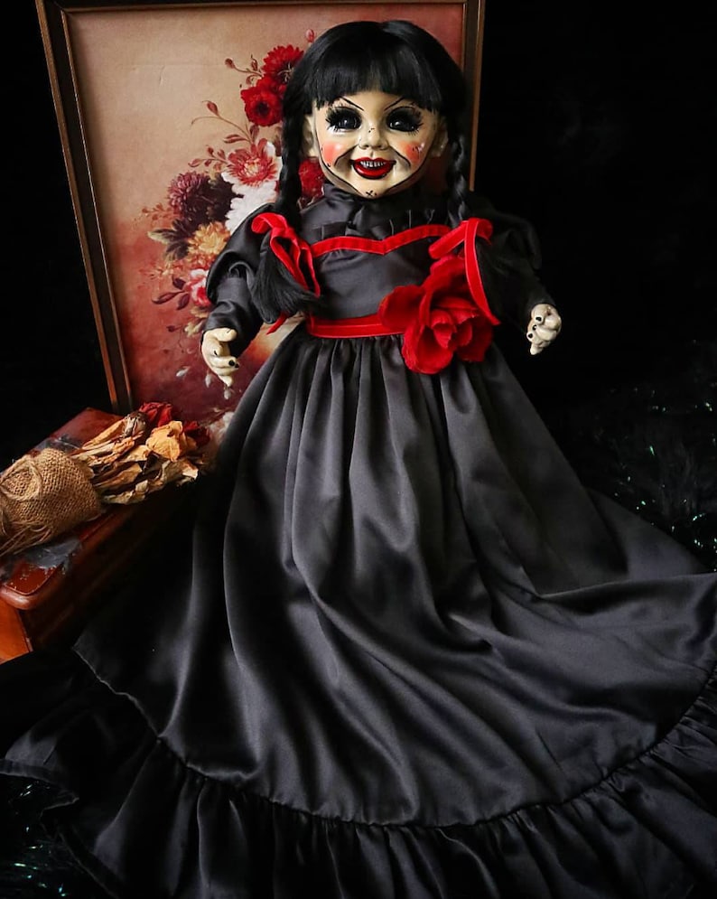 May include: A vintage-style doll with a porcelain face, black hair in pigtails, and a black dress with red accents. The doll has a painted smile and is posed in front of a floral print.