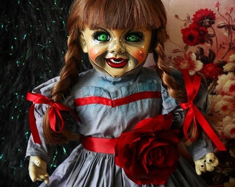 May include: A creepy porcelain doll with red hair in pigtails, wearing a blue and red dress with a red rose on the front. The doll has green eyes and a wide, toothy smile.