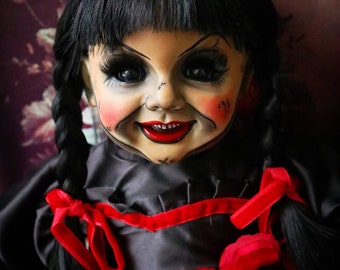 May include: A vintage doll with black hair styled in braids and bangs. The doll has a painted face with red cheeks and lips, and large black eyes. It wears a black dress with red ribbon accents and a large red rose.