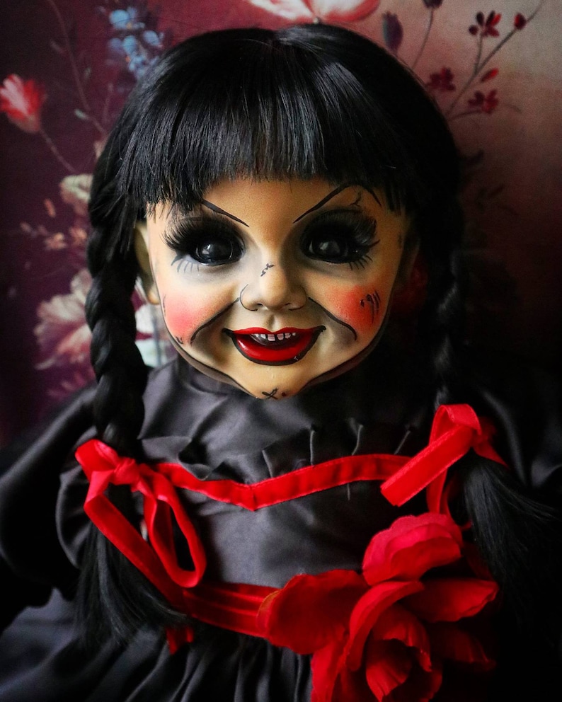 May include: A vintage doll with black hair styled in braids and bangs. The doll has a painted face with red cheeks and lips, and large black eyes. It wears a black dress with red ribbon accents and a large red rose.