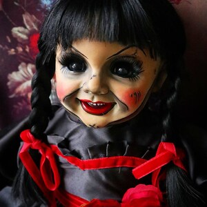May include: A vintage doll with black hair styled in pigtails, adorned with red ribbons. The doll has a pale face with rosy cheeks, dark eyes, and a sinister smile. It wears a black dress with a red rose accent.