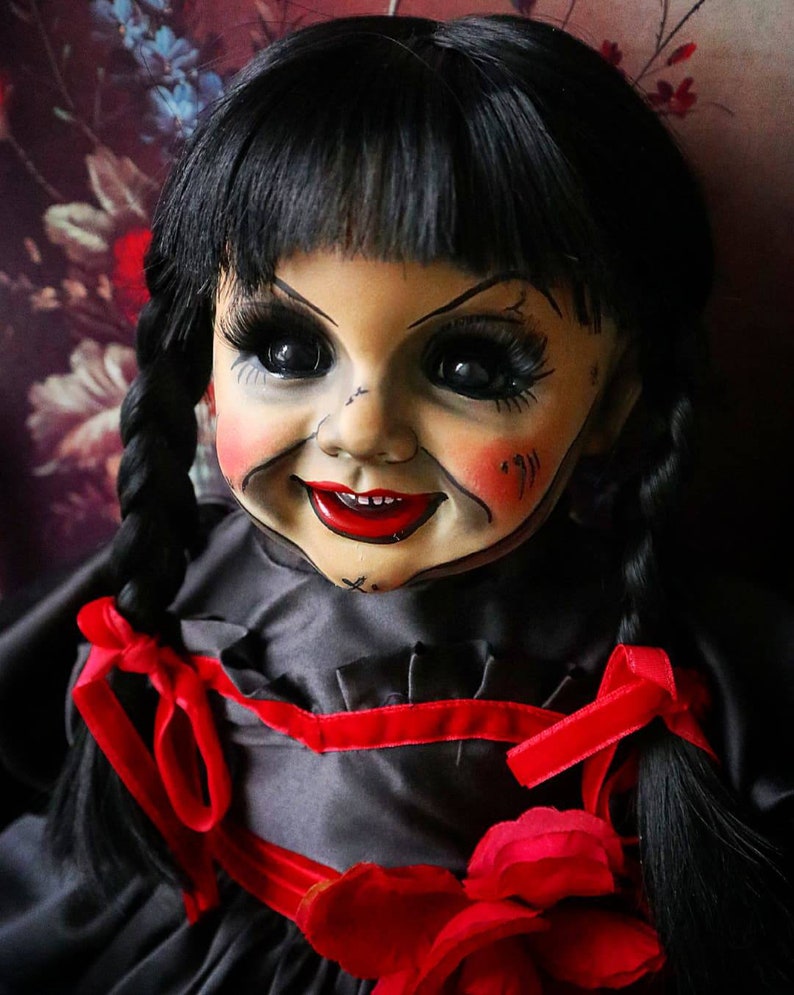 May include: A vintage doll with black hair styled in pigtails, adorned with red ribbons. The doll has a pale face with rosy cheeks, dark eyes, and a sinister smile. It wears a black dress with a red rose accent.