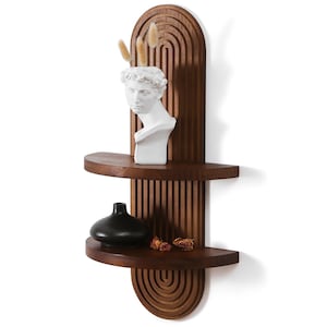 May include: A dark brown wooden wall shelf featuring a vertical arched back panel with carved parallel lines. The shelf has two semi-circular tiers. The top tier holds a white bust sculpture with dried grass, and the bottom tier holds a black vase.