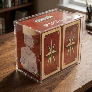 Puede incluir: Una vitrina de acrílico transparente que contiene un set de caja completa de Delicious in Dungeon. La caja presenta una cubierta roja con texto blanco, la ilustración de un personaje de pelo corto y dos paneles decorados con grandes estrellas brújula doradas.