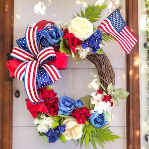 May include: A patriotic wreath with red, white, and blue flowers and a bow, accented with an American flag. The wreath is made of woven branches and is displayed on a wooden door.
