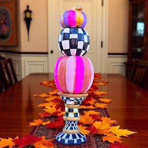 May include: A decorative pumpkin-themed centerpiece with three stacked, colorful pumpkins. The top pumpkin is purple, orange, and yellow. The middle pumpkin is black and white checkered. The bottom pumpkin is pink and red striped. The centerpiece sits on a patterned stand and is surrounded by fall leaves.